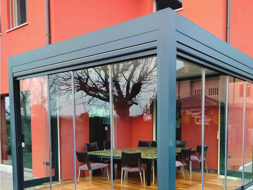 Pergola grigia chiusa con Sliding Floor davanti a una casa rossa, con il tavolo da pranzo dentro